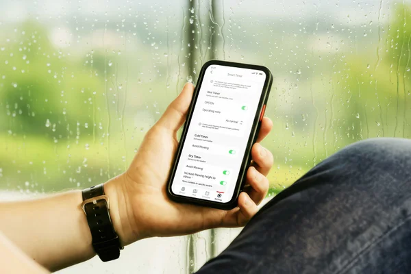 Hand holds phone by rainy window, smart timers adjust mowing for wet weather.